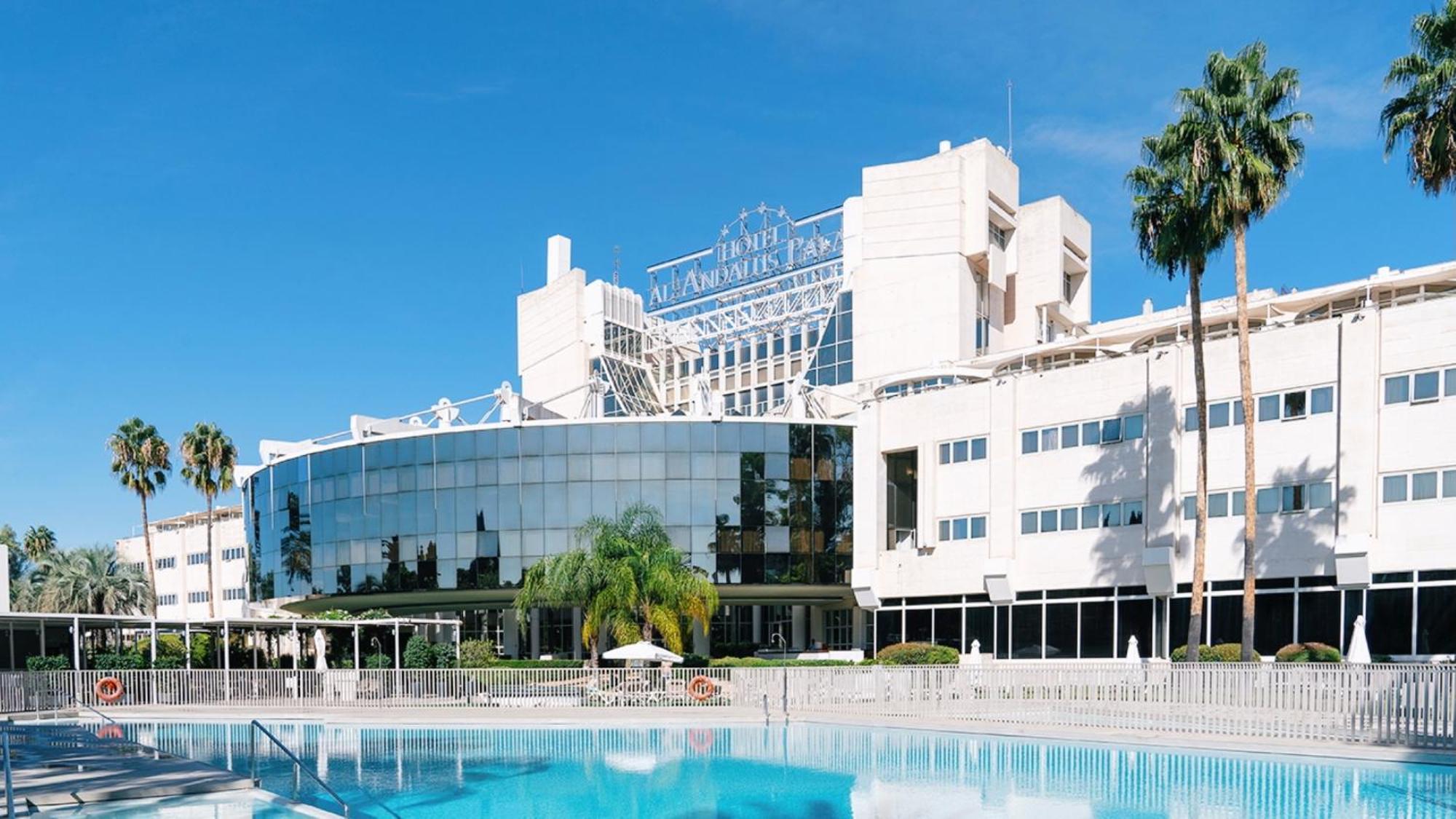 Silken Al-Andalus Palace Sevilla Exterior foto The photo shows a modern hotel building with a large glass facade, reflecting the surrounding area. There are palm trees in the foreground, and a swimming pool in the forefront of the image. The hotel has a stylish design, with a prominent sign on to